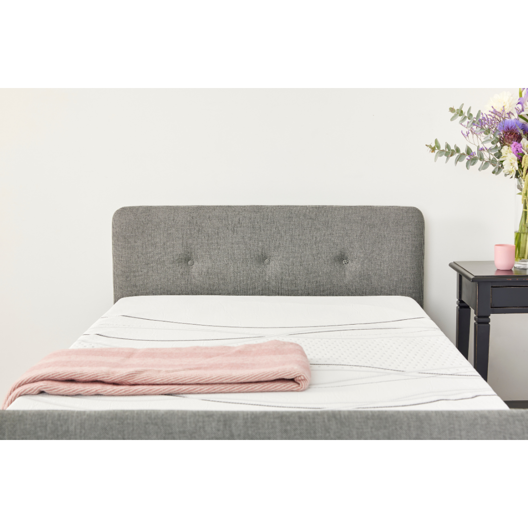 A neatly made bed with an iCare Medical Headboard and Footboards for iCare Beds, a pink blanket at the foot, and a black nightstand with a vase of colorful flowers and a pink cup beside it against a plain white wall.