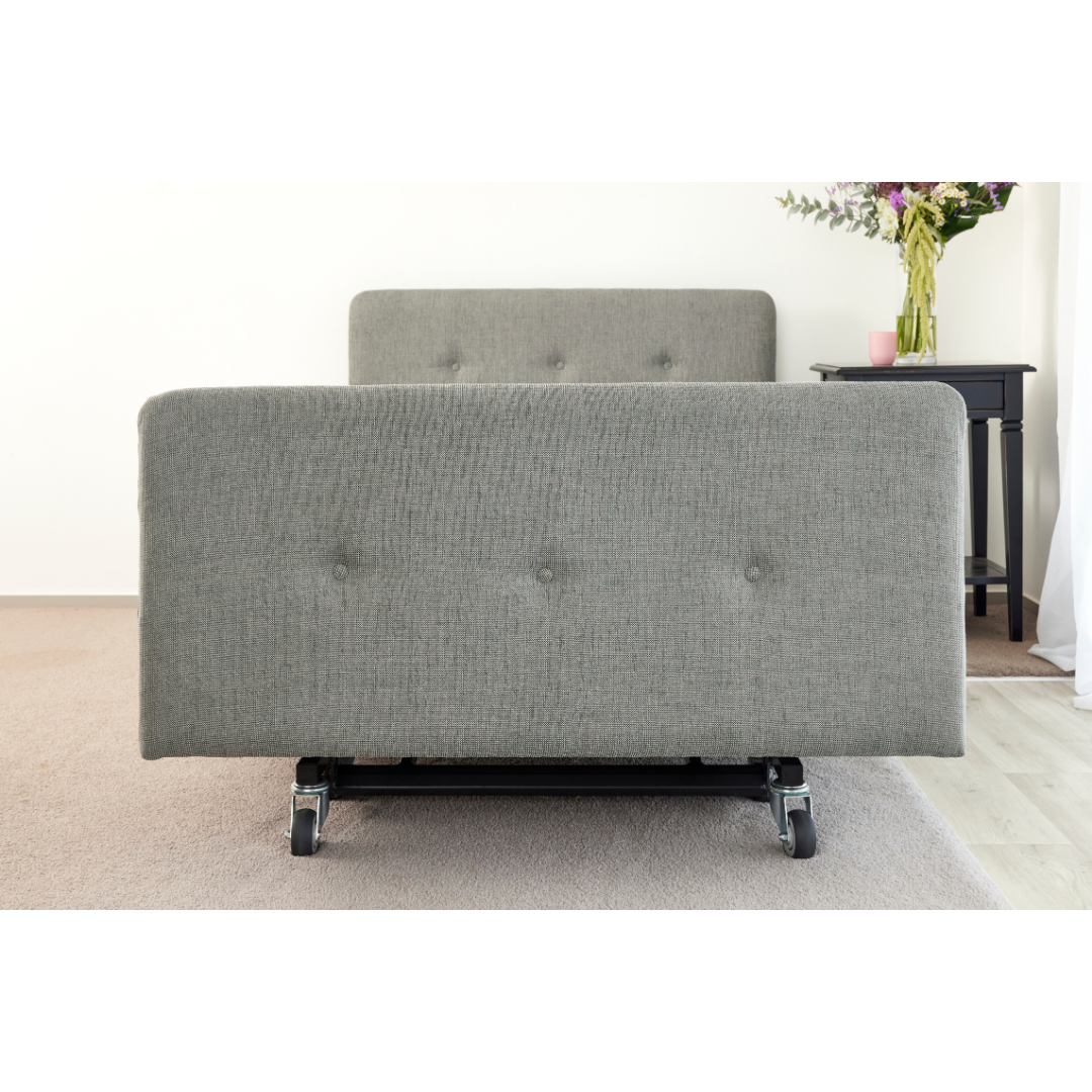 A gray iCare Medical Headboard and Footboards set for iCare Beds on wheels sits on carpet near a black bedside table with a vase, pink cup, and book, combining style with improved bed aesthetics.