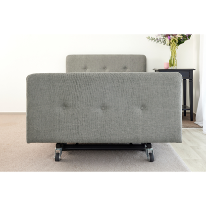 A gray iCare Medical Headboard and Footboards set for iCare Beds on wheels sits on carpet near a black bedside table with a vase, pink cup, and book, combining style with improved bed aesthetics.