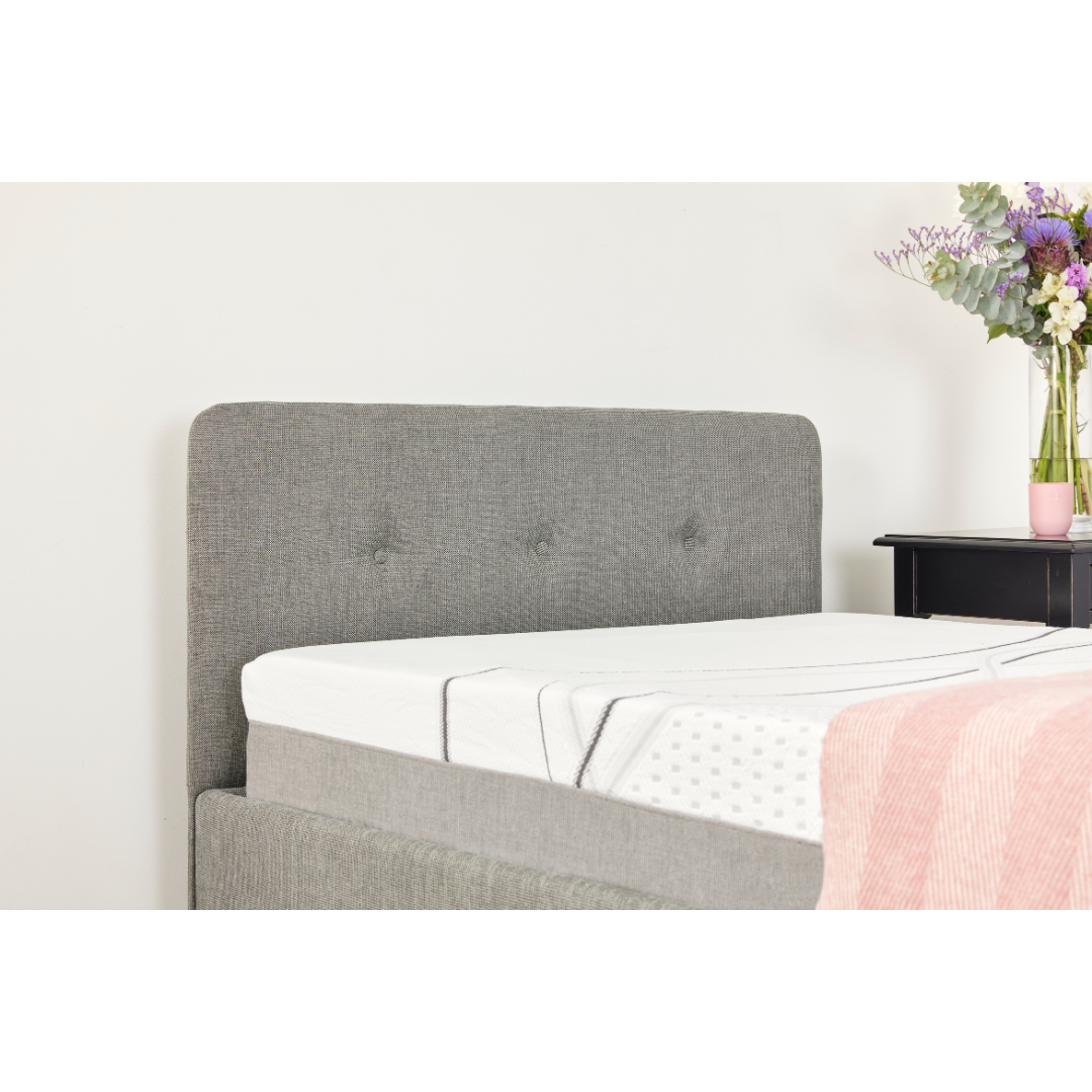 The iCare Medical Headboard and Footboards for iCare Beds provide a modern upgrade with a tufted grey headboard, paired with a white mattress and pink blanket. A black nightstand and floral arrangement complete the chic look against a plain white wall.