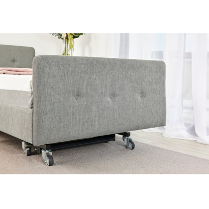 A close-up of the iCare Medical Headboard and Footboards for iCare Beds, shown in grey upholstery with wheels on a light carpet, near a window with sheer curtains and a vase of flowers in the background.