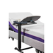 An adjustable hospital bed with a height-adjustable backrest features an iCare Medical Over Bed Tilting Table holding a tray and a white mug. The bed has a white mattress with purple accents and a light gray frame.