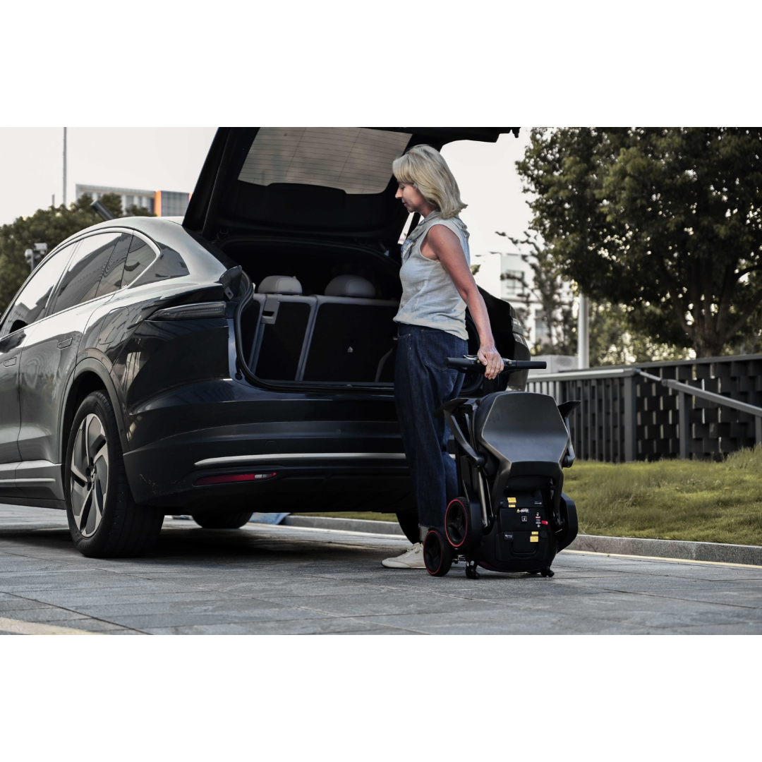 A woman stands by an open car trunk, holding a folded iMOVE AirCarbon Folding Carbon Fiber Scooter.