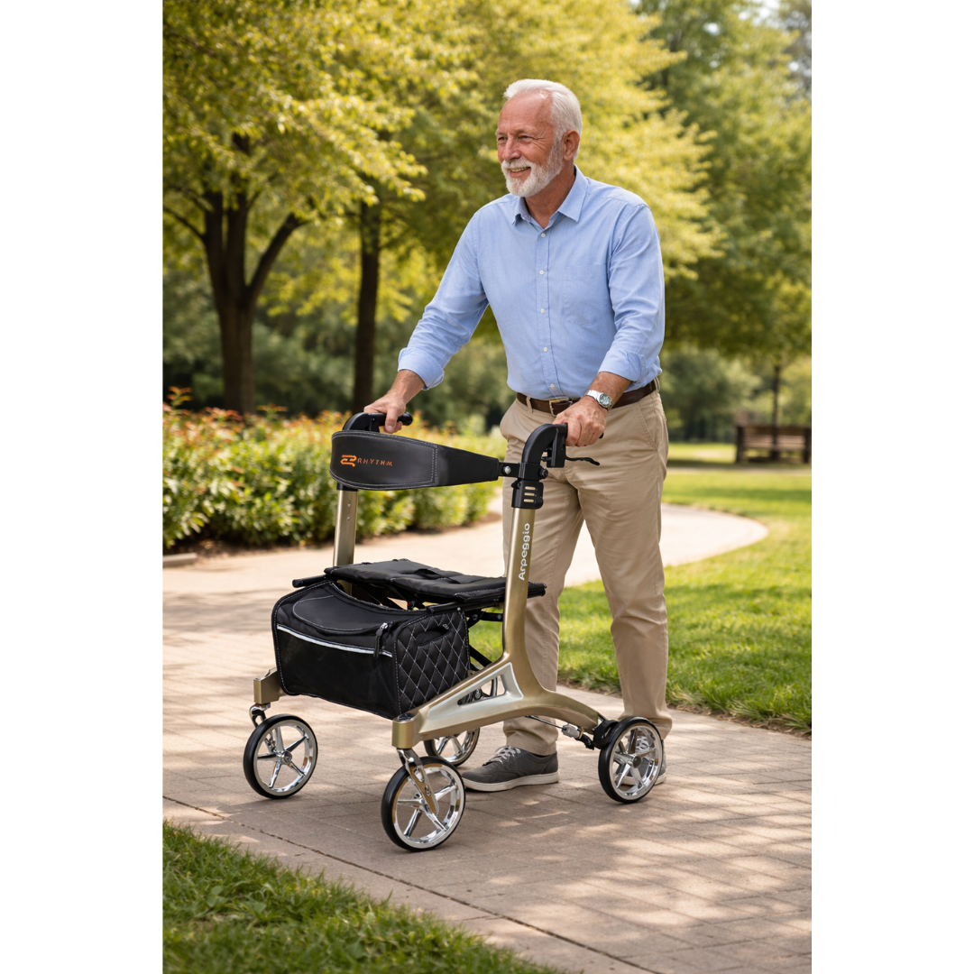 An older man with gray hair and a beard smiles while walking outside on a paved path using the Rhythm Arpeggio Folding Portable Rollator with XL 8" Wheels in Champagne, which features a black storage bag. Green trees and shrubs are behind him.