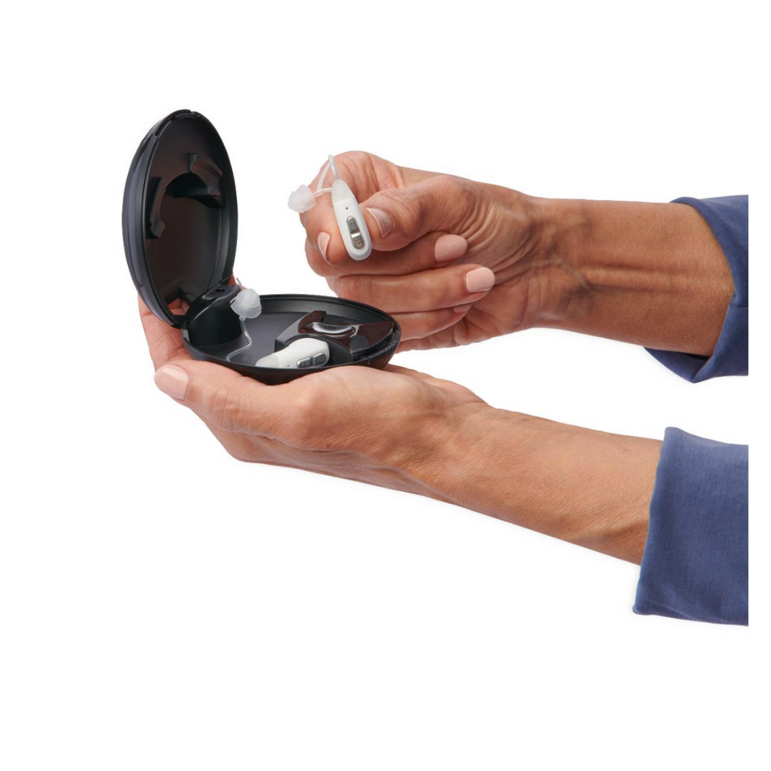 A person in a blue long-sleeve shirt holds a white Medline Rechargeable Digital OTC Hearing Aid (Behind-The-Ear style) with volume control above an open black charging case containing another aid.