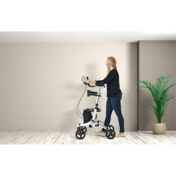 A person with a Medline Upright Aluminum Rollator, featuring ergonomic handbrakes, is in a room with wooden flooring and a potted plant. Wearing a mask, black top, and jeans, they move rightward against a plain beige wall.