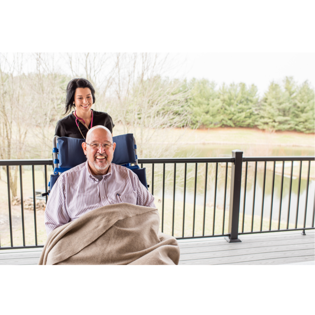 An older man smiles from a Med-Mizer FLEXTILT Reclining Transfer Chair, covered with a blanket, as a caregiver stands behind him on a porch overlooking trees and a pond.