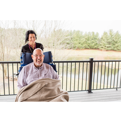 An older man smiles from a Med-Mizer FLEXTILT Reclining Transfer Chair, covered with a blanket, as a caregiver stands behind him on a porch overlooking trees and a pond.