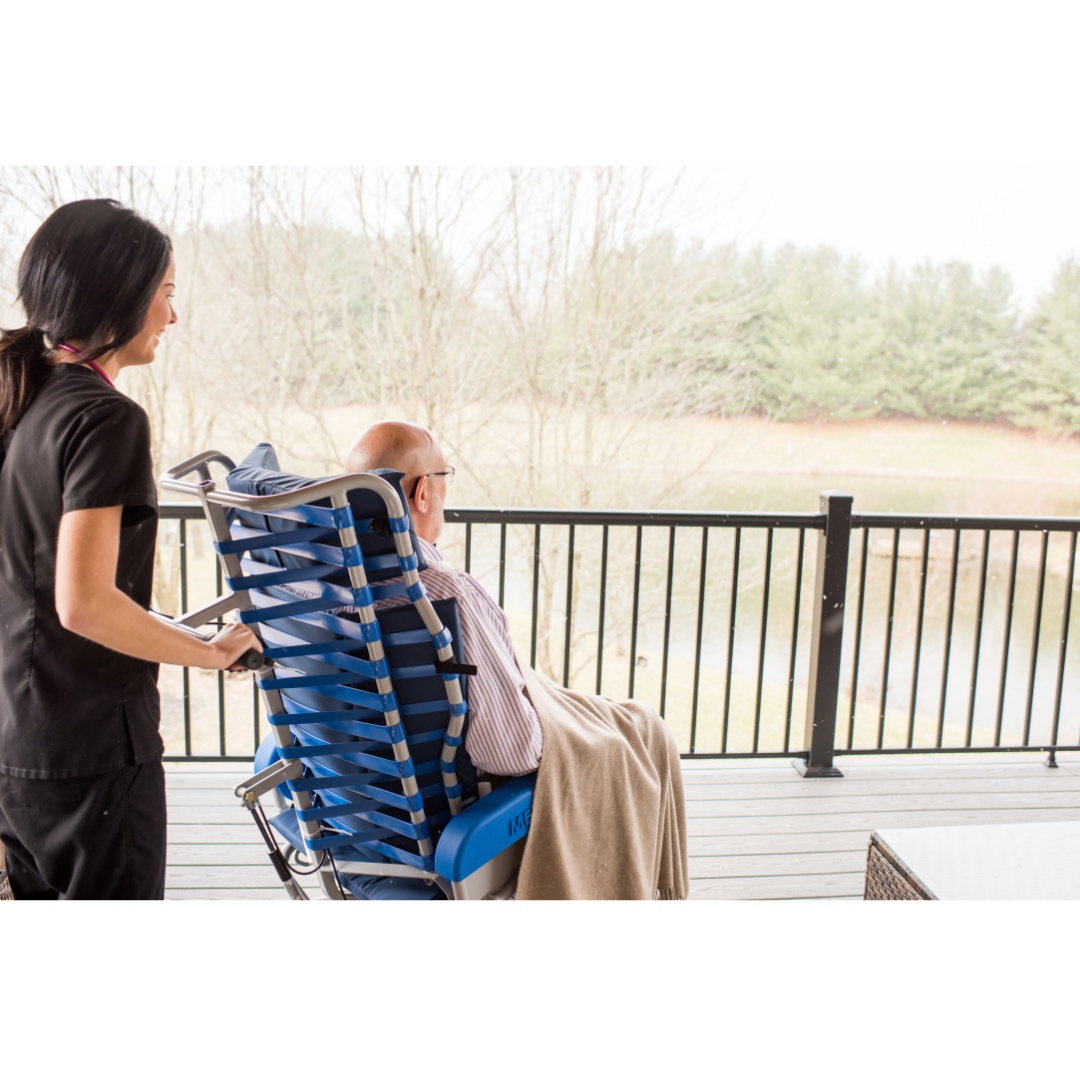 A caregiver stands behind an elderly man seated in a Med-Mizer FLEXTILT Reclining Transfer Chair on a deck, overlooking trees and a pond. The man, covered with a beige blanket, enjoys mobility support and comfort.