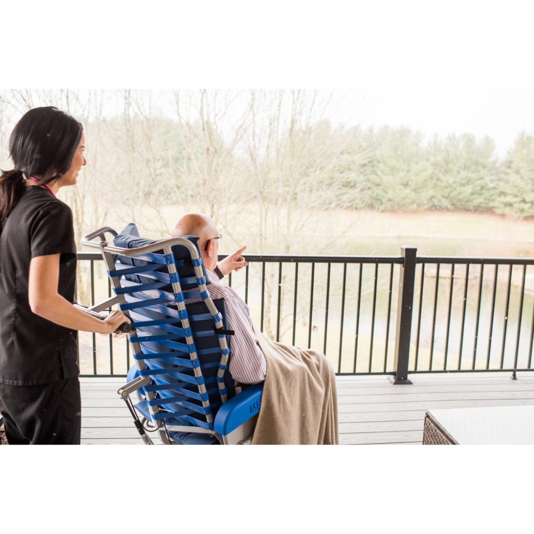 A caregiver stands by an elderly man seated in the Med-Mizer FLEXTILT Reclining Transfer Chair, covered with a blanket, as he points out toward a scenic pond and trees from a wooden deck.