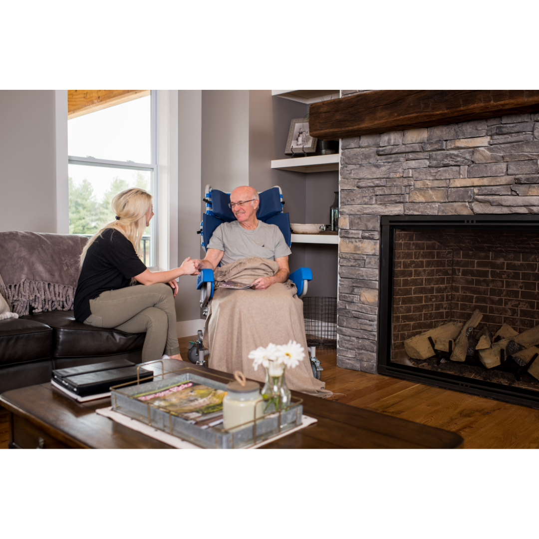A woman sits on a couch, holding hands with an older man covered by a blanket in the cozy living room. He is seated in a Med-Mizer FLEXTILT Reclining Transfer Chair near the fireplace and large window.