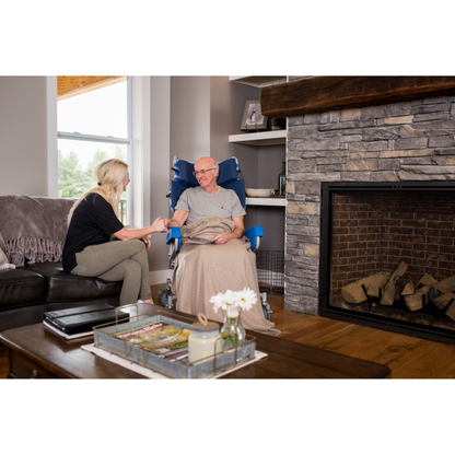 A woman sits on a couch, holding hands with an older man covered by a blanket in the cozy living room. He is seated in a Med-Mizer FLEXTILT Reclining Transfer Chair near the fireplace and large window.