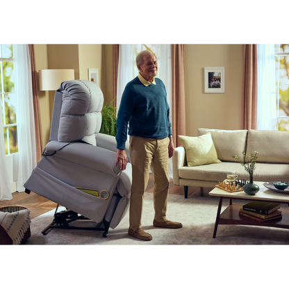 An elderly man stands next to a Journey Perfect Sleep Reclining Lift Chair with Heat in a cozy living room. The chair, partially lifted, offers therapeutic heat and massage. The space has a beige couch, decor on the coffee table, and curtains on the windows. He wears a blue sweater and khaki pants.