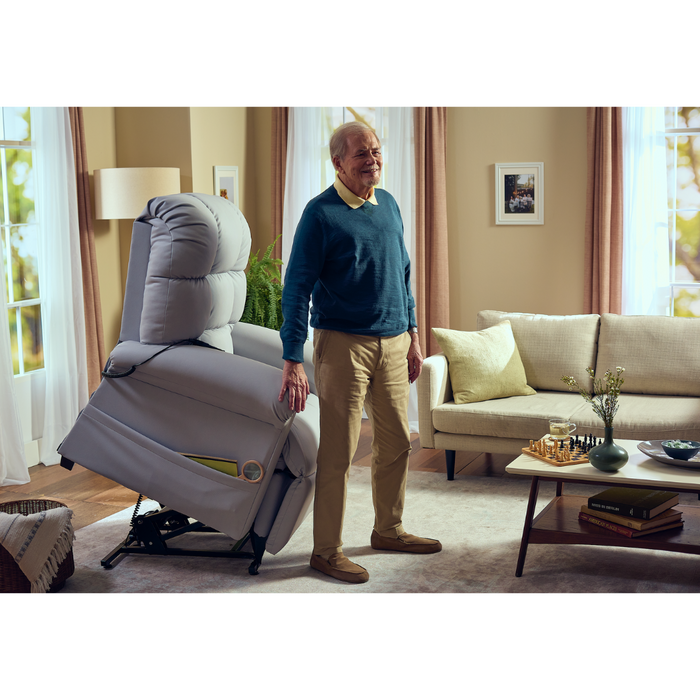 An elderly man stands next to a Journey Perfect Sleep Recliner & Lift Chair with Heat in a snug living room. Hes clad in a blue sweater and beige pants, while the room features a beige sofa, framed pictures, and a small table with books and a wine glass.