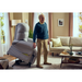 An elderly man stands next to a Journey Perfect Sleep Recliner & Lift Chair with Heat in a snug living room. Hes clad in a blue sweater and beige pants, while the room features a beige sofa, framed pictures, and a small table with books and a wine glass.