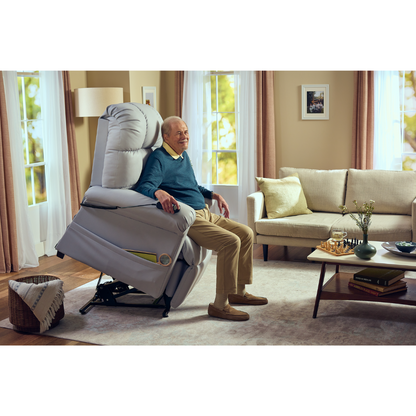 An elderly man relaxes in the Journey Perfect Sleep Reclining Lift Chair with Heat, partially elevated for comfort. The room is inviting with a cozy sofa, coffee table, and gentle lighting. Large windows showcase trees outside, amplifying the welcoming ambiance.