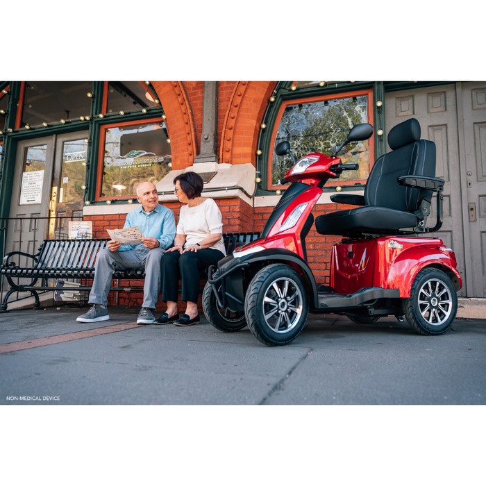 An older man and woman sit on a bench outside a brick building, looking at a brochure together. Nearby, a Pride Baja® Raptor® 2 Bariatric 4-Wheel Scooter with LED lighting is parked, offering stylish outdoor mobility.