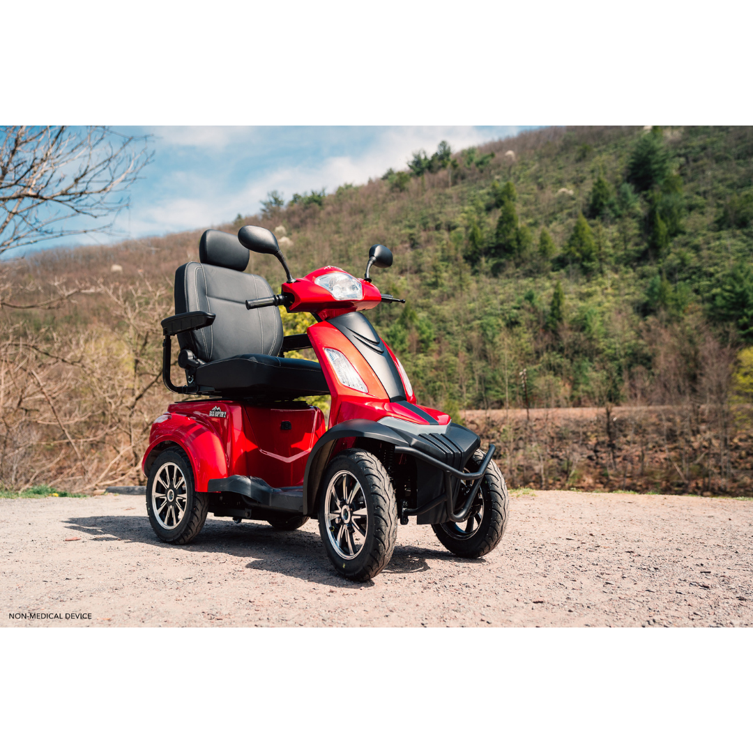 A Pride Baja® Raptor® 2 Bariatric 4-Wheel Scooter in red, featuring two black seats and LED lighting, is parked on a gravel path outdoors among green hills and trees beneath a partly cloudy sky.