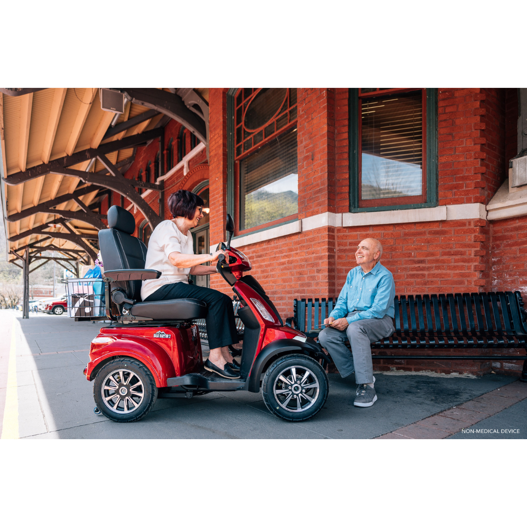 A woman enjoys premium comfort on a red Pride Baja® Raptor® 2 Bariatric 4-Wheel Scooter while chatting with an elderly man on a bench outside a brick building, showcasing the scooters outdoor mobility.