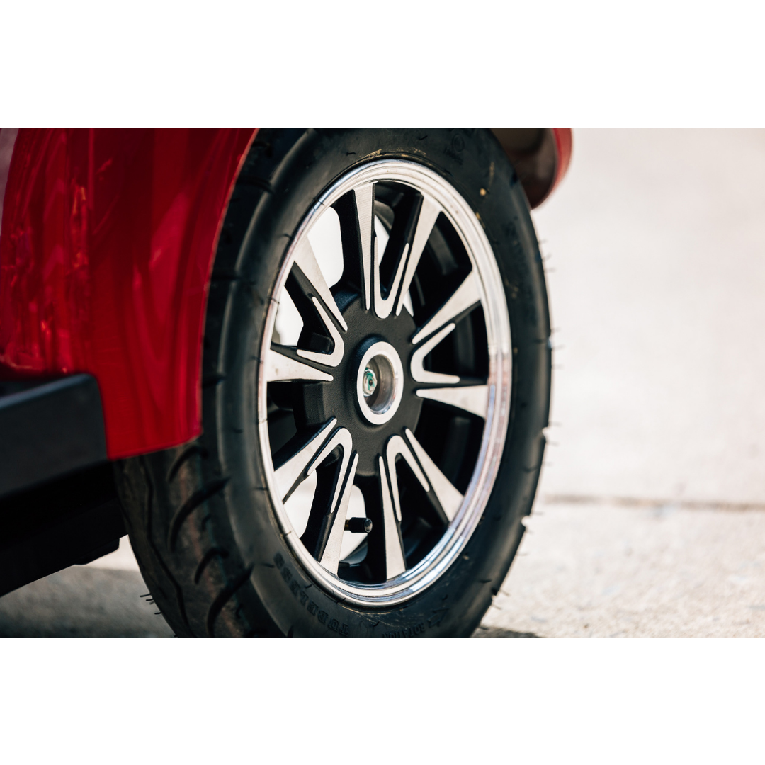 Close-up of a black tire with a silver and black alloy wheel on a red Pride Baja® Raptor® 2 Bariatric 4-Wheel Scooter, parked on concrete. The image highlights the premium design, comfort, and outdoor mobility features of the scooter.