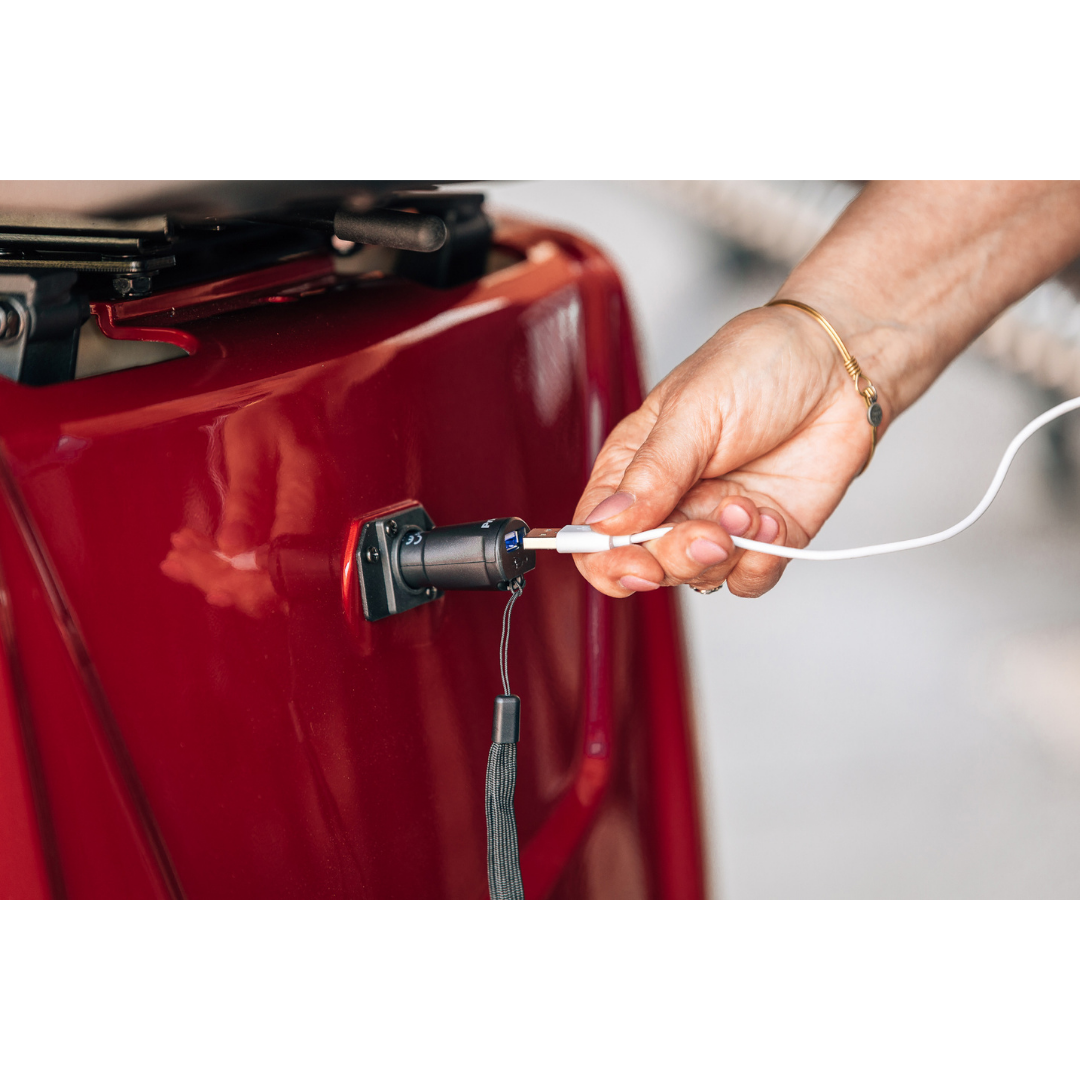 A hand plugs a white charging cable into the port of a shiny red Pride Baja® Raptor® 2 Bariatric 4-Wheel Scooter, showcasing its premium comfort and advanced outdoor mobility.