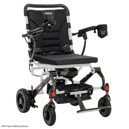 The Pride Mobility Jazzy® Carbon Ultralite Powerchair is a black and silver travel chair with cushioned seat, armrests, footrest, small front wheels, larger rear wheels, and a detachable joystick with USB charger. It includes a mesh cup holder and is labeled FDA Class II Medical Device.