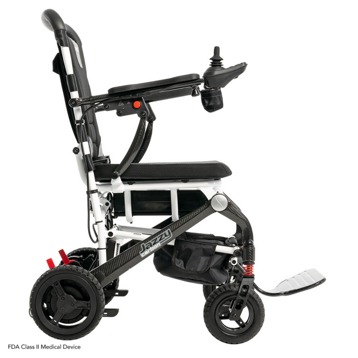 A side view of the Pride Mobility Jazzy® Carbon Ultralite Powerchair shows a black and white ultra-lightweight design with a removable joystick featuring a USB charger on the right armrest. It has four wheels, a padded seat and backrest, footplate, airline-compliant lithium battery, and an elegant Jazzy frame logo.