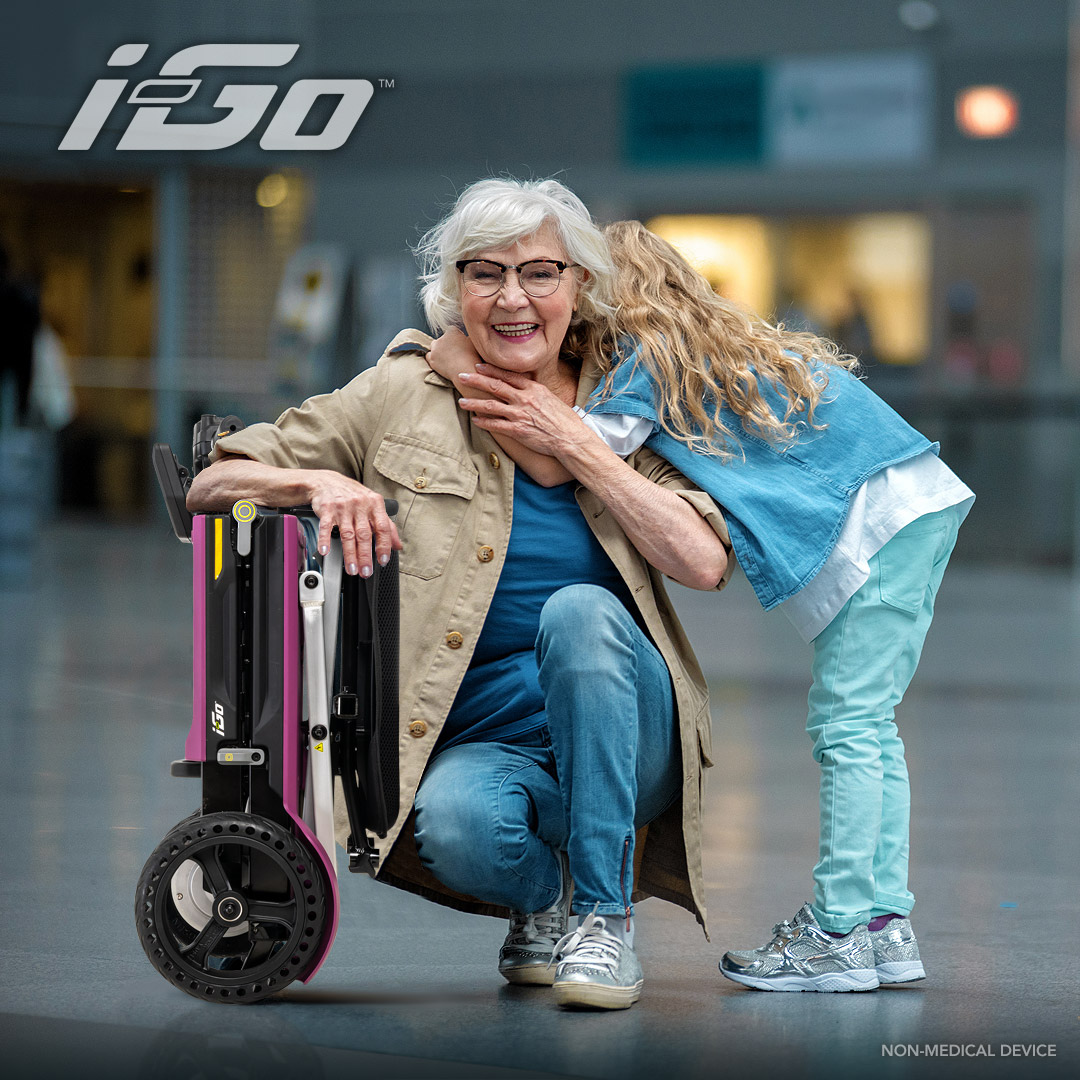 An elderly woman in a light jacket and glasses kneels beside a Pride Mobility i-Go™ Portable Travel Scooter. A young blonde girl hugs her from behind as they smile in a bright, open indoor space.