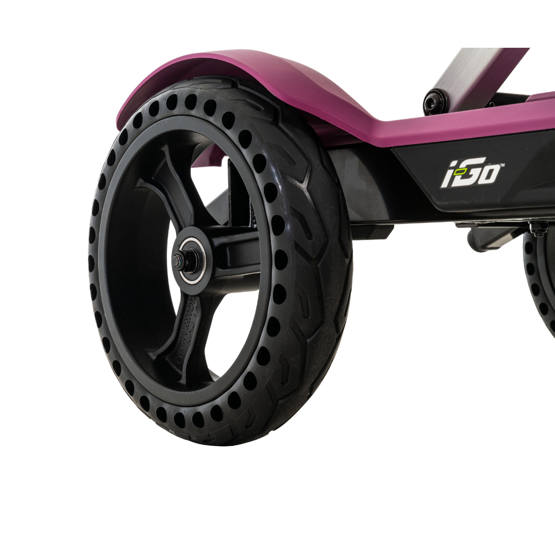 A close-up of the purple Pride Mobility i-Go™ Portable Travel Scooter highlights its rear wheel and suspension system. The sleek scooter features a black, perforated tire on a silver metal frame with an Ultra-Compact Profile and i3O branding on the side.