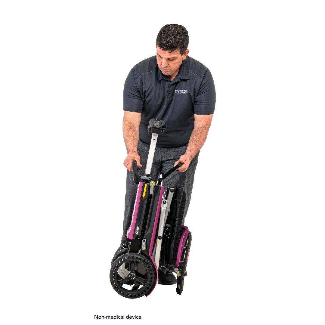 Someone in a dark polo and gray pants demonstrates folding the Pride Mobility i-Go™ Portable Travel Scooter with large wheels, showcasing its ultra-compact profile and functional handle. The text Non-medical device appears at the bottom.
