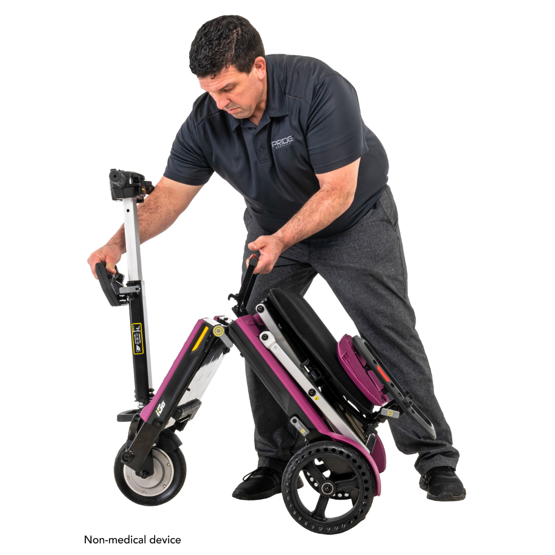 A man demonstrates the Pride Mobility i-Go™ Portable Travel Scooter, a mobility device with a purple frame and Feather-Touch Disassembly for easy folding. He wears a dark polo shirt and gray pants, standing against a plain white background.