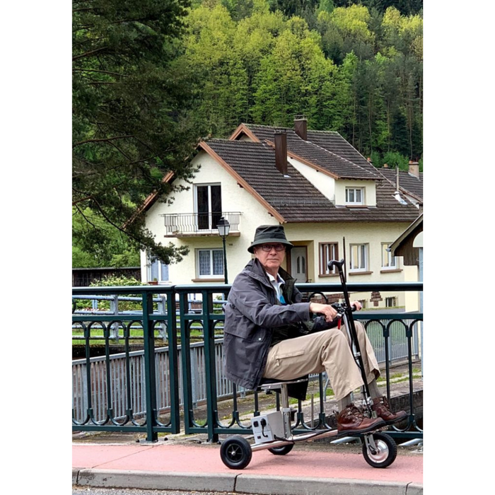 An older adult, wearing a hat and jacket, travels across a bridge on the SmartScoot® Ultra Light Mobility Scooter—FAA Approved. A house surrounded by lush greenery creates a picturesque backdrop.