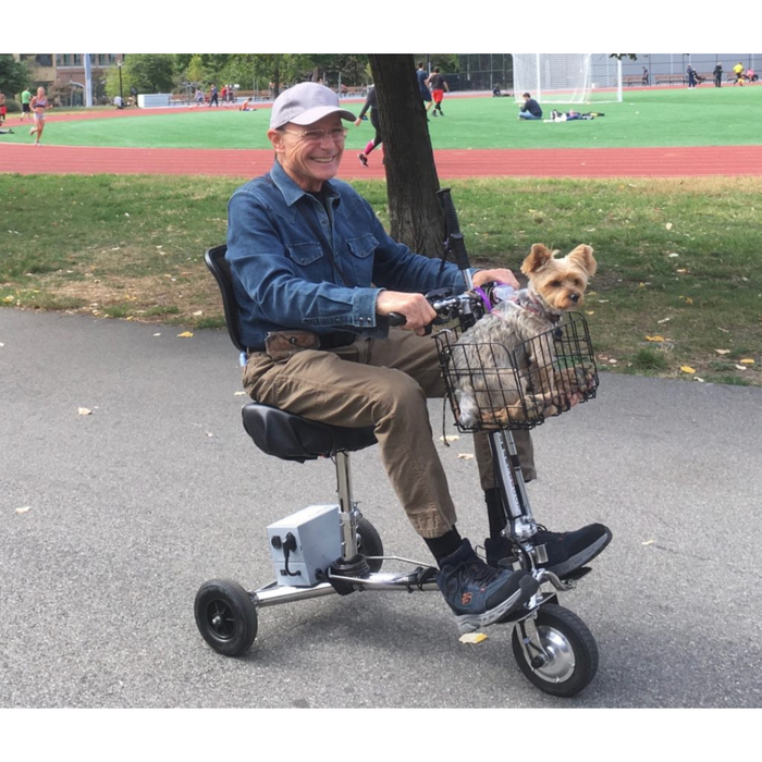 An elderly man in a cap and denim jacket rides his SmartScoot® Ultra Light Mobility Scooter with FAA approval. His small dog is snug in the front basket as they cruise along a path near a lively sports field, while he considers his next travel adventure.