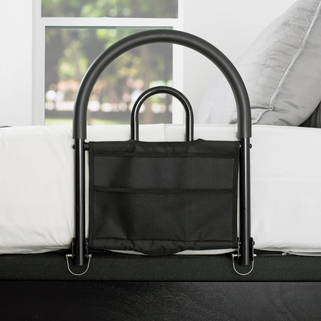 The Stander Bed Rail Advantage Traveler, a portable black safety rail with a storage pouch for traditional beds, is attached to the bedside; outside the nearby window is a green outdoor scene.