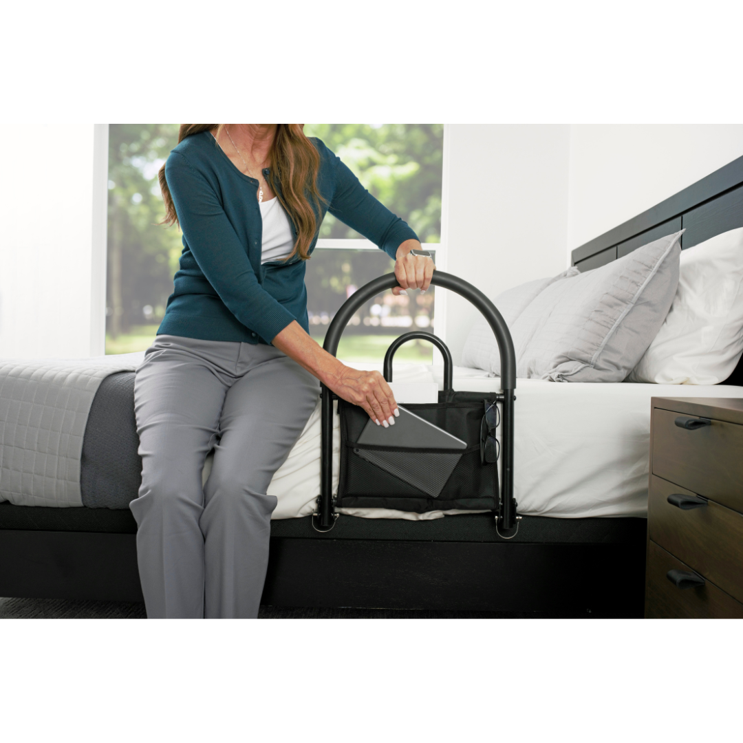 A woman sits on a bed using the Stander Bed Rail Advantage Traveler, a black portable bed safety rail with an attached storage pouch, while holding a tablet in her bright, modern bedroom.