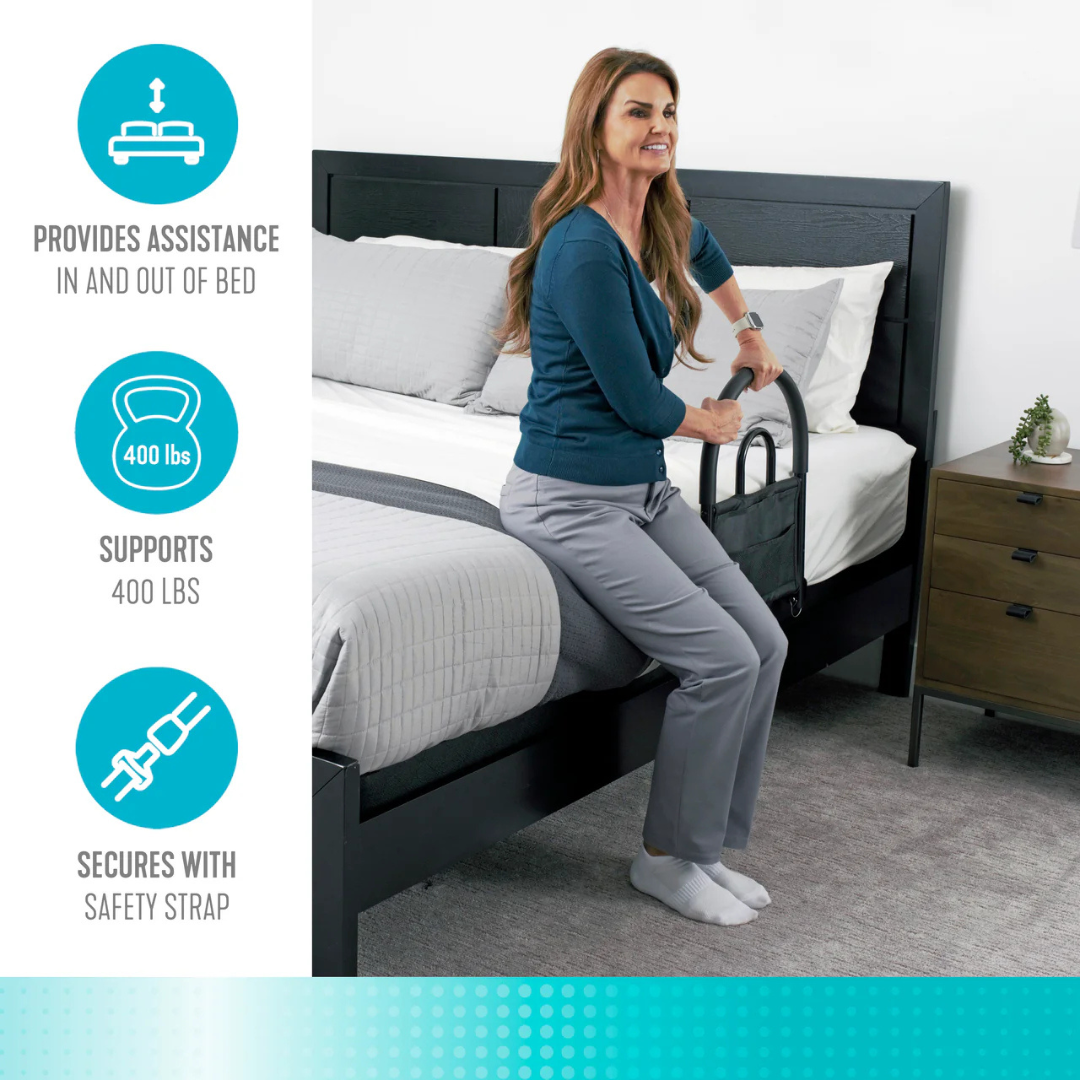 A woman uses the Stander Bed Rail Advantage Traveler, a black portable safety rail, while sitting on a made bed. Graphics show its bariatric support handle assists users, supports up to 400 lbs, and secures with a safety strap.