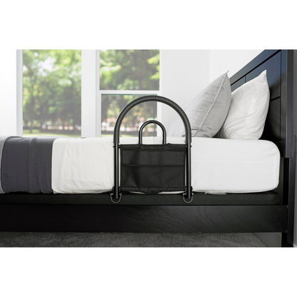 The Stander Bed Rail Advantage Traveler, a portable bed safety rail with a storage pouch, is attached to the side of a modern black bed with white bedding and gray pillows, positioned by large windows overlooking greenery.