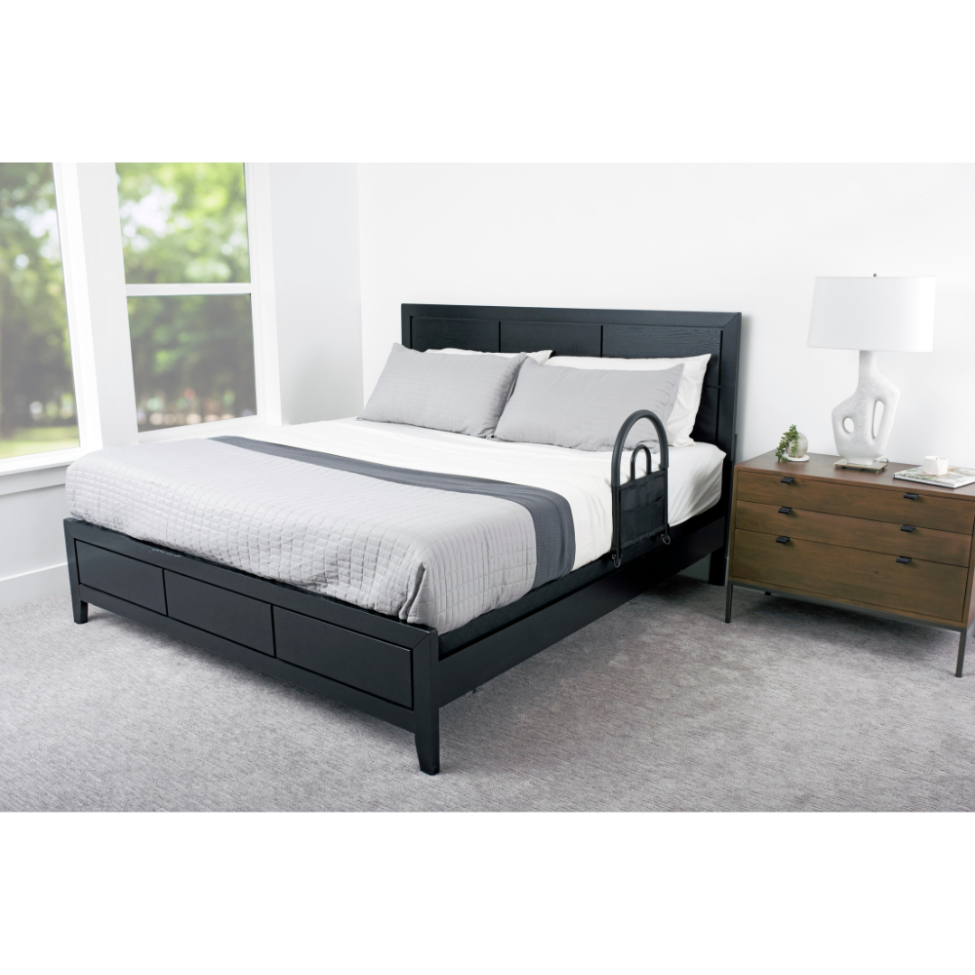A modern bedroom features a black bed frame, gray and white bedding, the Stander Bed Rail Advantage Traveler for safety on the side, a wooden nightstand with a lamp and decor, and large windows allowing in natural light.