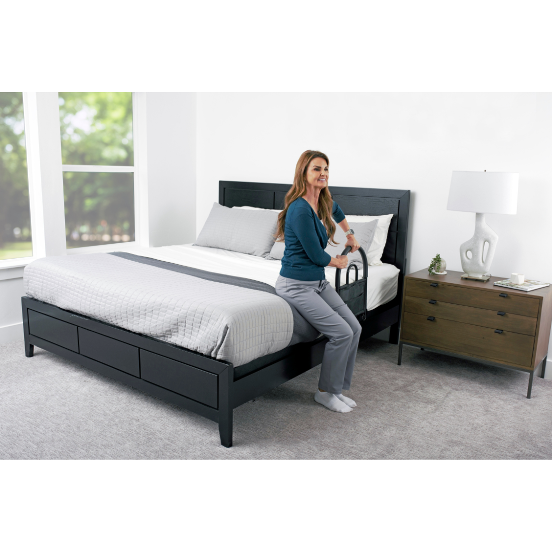 A woman sits on the edge of a neatly made bed, holding the Stander Bed Rail Advantage Traveler – Portable Bed Safety Rail for support. The bright room features large windows, a nightstand, and a lamp beside the bed.