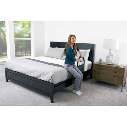 A woman sits on the edge of a neatly made bed, holding the Stander Bed Rail Advantage Traveler – Portable Bed Safety Rail for support. The bright room features large windows, a nightstand, and a lamp beside the bed.
