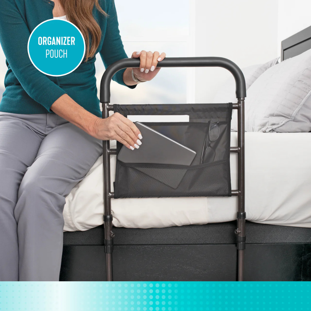 A woman sits on a bed, placing a tablet into a black organizer pouch attached to the Stander Stable Rail - Bedside Standing Aid with Extendable Legs, which provides sturdy support and convenient storage. A label above reads ORGANIZER POUCH.