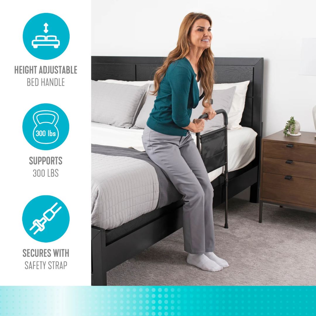 A woman uses the Stander Stable Rail - Bedside Standing Aid with Extendable Legs, a black bed handle attached to the bed for support when standing. It is height adjustable, supports 300 lbs, has a safety strap, and is portable.