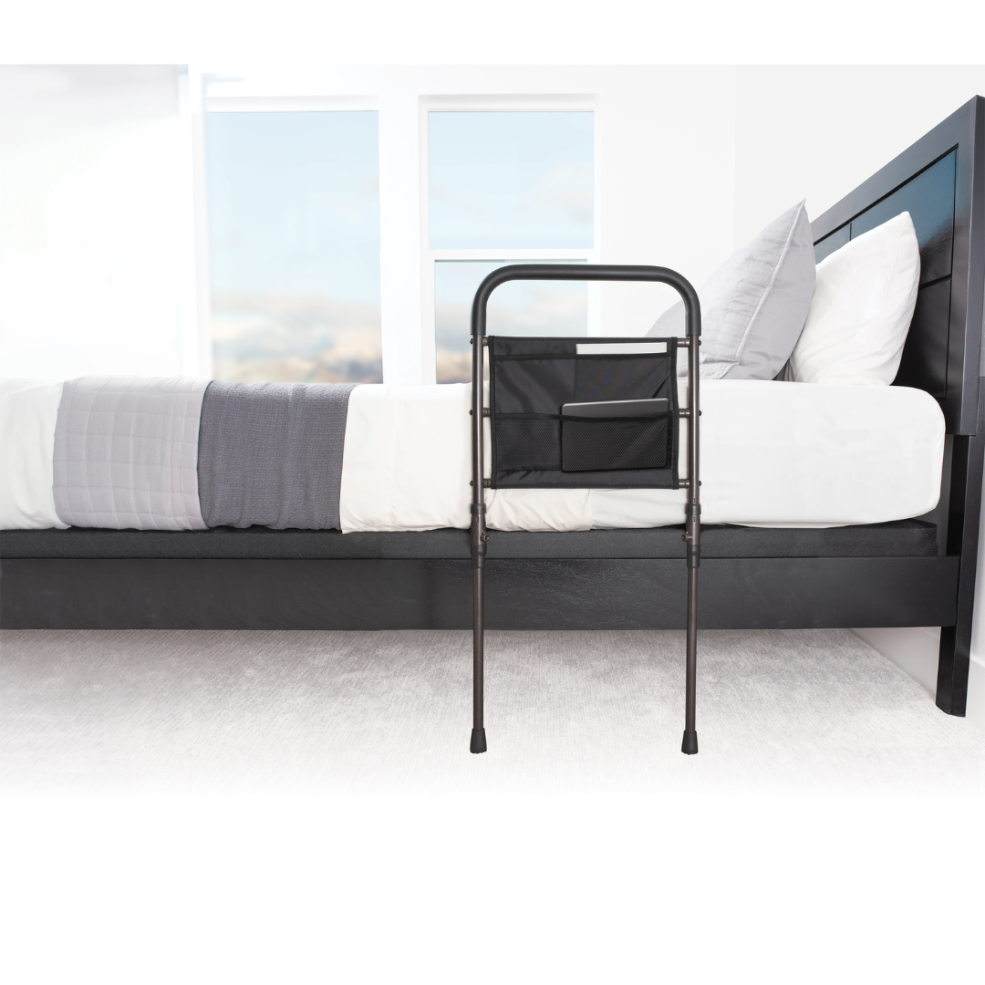 The Stander Stable Rail - Bedside Standing Aid with Extendable Legs is secured to the side of a modern bed with white and gray bedding in a bright, carpeted bedroom.