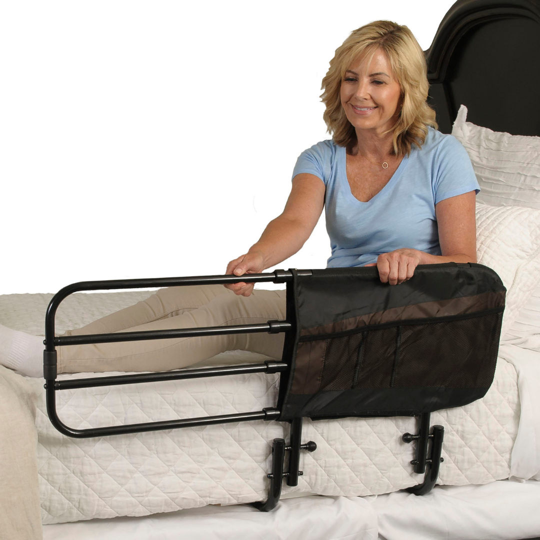 A woman sits on a bed, holding the Stander EZ Adjust Bed Rail with mesh pouch, attached to the bed for support, safety, and fall protection.