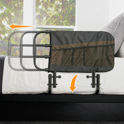 The Stander EZ Adjust Bed Rail in black and gray attaches to an adult bed, features mesh storage pockets, slides horizontally, folds down for easy access, and provides fall protection. A window with green trees is visible behind the bed rail.