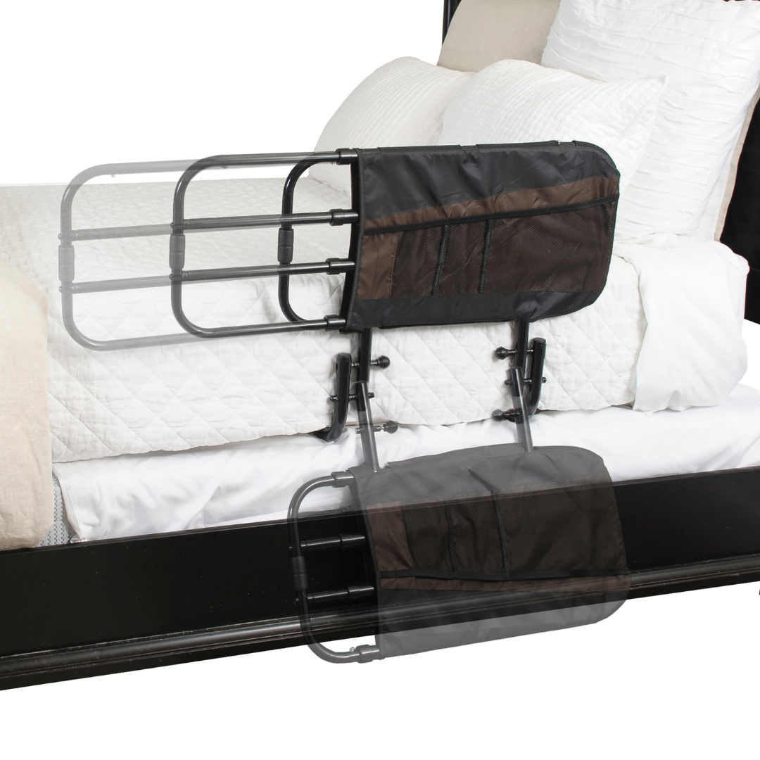 The Stander EZ Adjust Bed Rail is shown upright and folded down, attached to a dark bed frame for fall protection. It includes a black mesh storage pouch and fits between white bedding and the frame.
