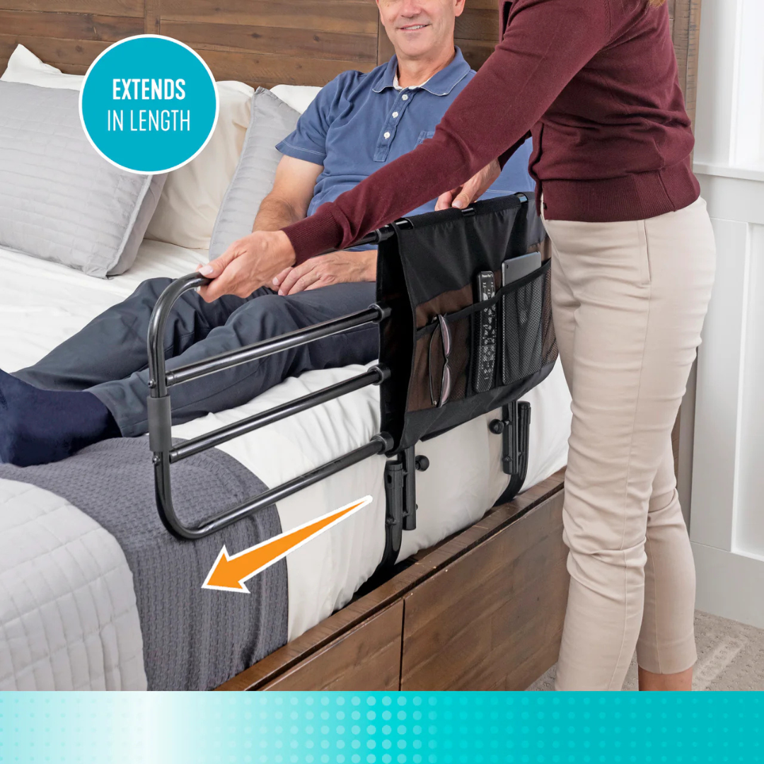 A person stands beside a bed, adjusting a Stander EZ Adjust Bed Rail for a seated man. The black rail, labeled EXTENDS IN LENGTH, features pockets for items and a remote, offering fall protection for adults.