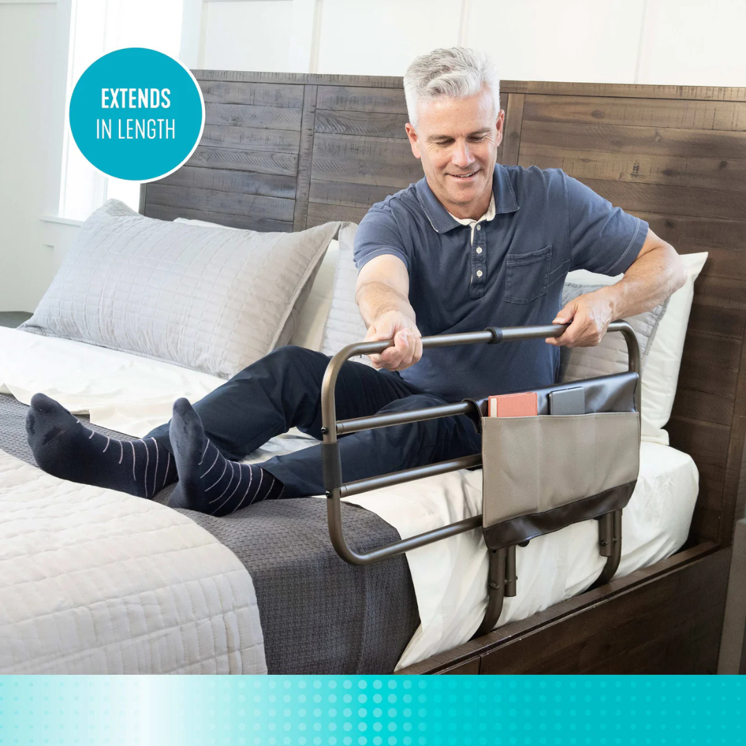 A smiling man sits on a bed with gray bedding and a wooden headboard, holding the Signature Life Sleep Safe Extendable Bed Rail with a storage pouch. The rail is extended and labeled EXTENDS IN LENGTH. See product specs for more details.