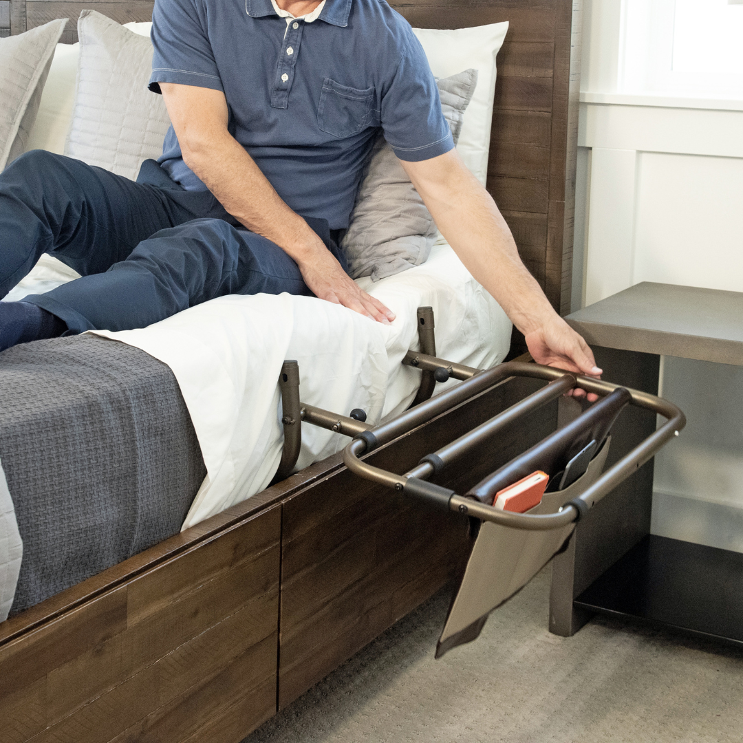 A person sitting on a bed reaches for the Signature Life Sleep Safe Extendable Bed Rail, which features an attached organizer holding a phone and tablet, next to a wooden nightstand. Warranty details and specs are available for peace of mind.