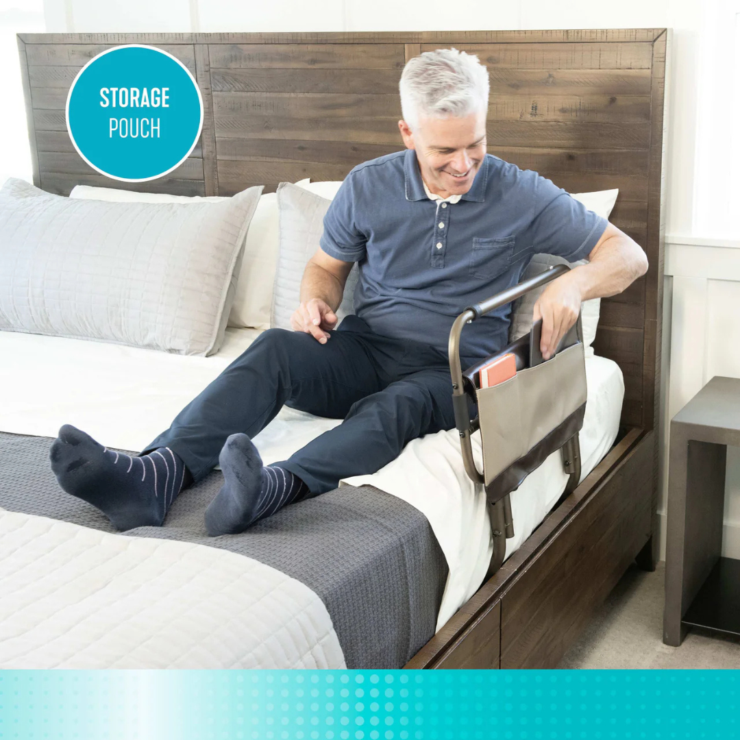 An older man sits on a bed, smiling as he places a book into a tan pouch attached to the Signature Life Sleep Safe Extendable Bed Rail. The well-lit room and cozy white-gray bedding create an ideal spot for reading or relaxing.